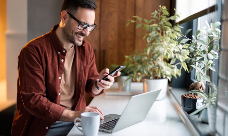 Satisfied modern millennial man in stylish casual clothes using smartphone and laptop computer for electronic banking, making reservation, online shopping and payments while spending time at home.