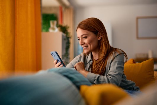 Smiling mid-adult woman relaxing on a cozy sofa at home, using her smartphone to connect with friends and enjoy her leisure time, embracing comfort and happiness in her evening routine